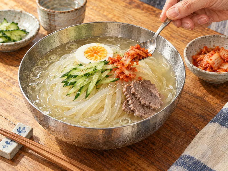 A hand adding a small spoonful of kimchi to adjust a bowl of chilled reimen